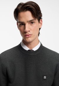 Dunkelgrauer Strickpullover mit strukturiertem Muster, rundem Halsauschnitt und einem kleinen schwarzen Logo-Patch auf der linken Brust. Wird über einem weißen Hemd mit Krawatte getragen.