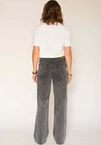 Jean flare taille haute en denim gris délavé, avec deux poches arrière et une finition subtilement texturée. Associé à une chemise blanche à manches courtes.