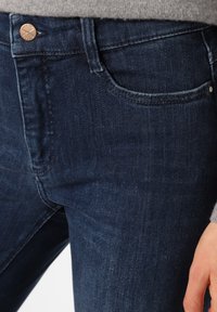 Jeans de denim de color azul oscuro con corte slim, que presentan un botón de cobre y un bolsillo frontal. La tela tiene una textura suave con un ligero desgaste.