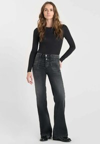 Camiseta negra de manga larga combinada con jeans oscuros de talle alto y acampanados, que cuentan con un cierre de botón y cinco bolsillos. Textura suave.