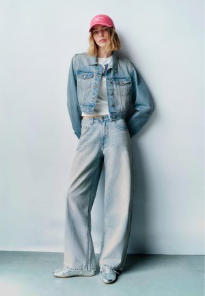 Fiatal nő rózsaszín sapkában, rövid farmerdzsekiben, fehér felsőben, bő szárú farmerral és tornacipőben, egy sima világos fal előtt állva.