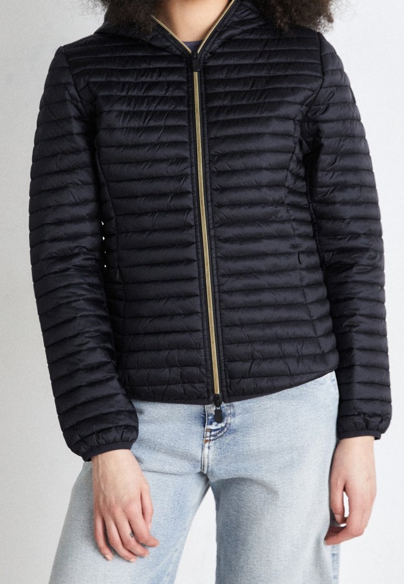 Personne portant une veste noire matelassée à fermeture éclair avec des surpiqûres horizontales et un jean bleu clair, les mains détendues le long du corps.