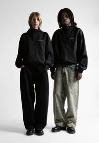 Két ember áll egymás mellett fekete cipzáras kapucnis dzsekikben, laza nadrágban és fekete cipőkben, egy sima fehér háttér előtt.