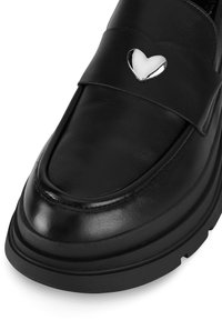Mocasín de cuero negro con un acabado suave, que presenta un acento en forma de corazón plateado en la correa y una suela gruesa negra con detalle texturizado.