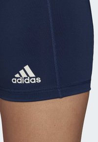 Námornícke modré športové šortky vyrobené z elastickej látky, s bielym logom Adidas, plochými švami a priliehavým strihom.