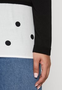 Maglietta nera a maniche lunghe con una parte inferiore bianca decorata con pois neri. Il tessuto appare morbido, con polsini a costine. Sotto viene mostrato del denim.