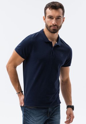Polo bleu marine à manches courtes, fabriqué en tissu doux. Comprend un col et un petit logo sur la poitrine gauche. Porté avec un jean bleu.