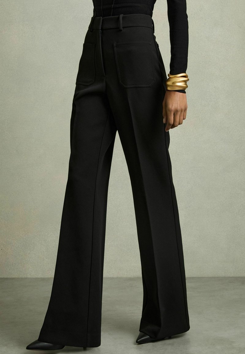 Pantalones de pierna ancha negros con una textura suave, cintura alta y dos bolsillos traseros. Combinados con tacones negros de punta y una pulsera de puño dorada.