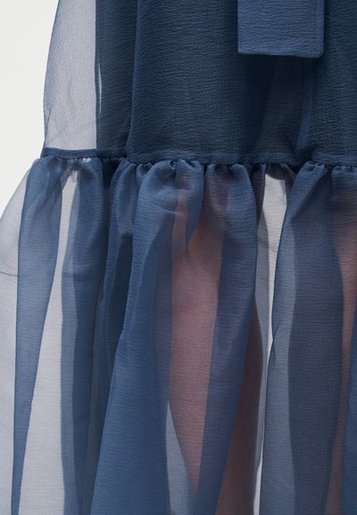 Διάφανο ύφασμα ναυτικού μπλε με λεπτομέρειες μαζεμένων βολάν πάνω σε υφασμένο φούστα, που αποκαλύπτει αχνές αποχρώσεις δέρματος μέσα από το διαφανές υλικό.