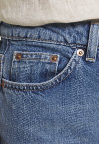 Denim-farkut keskipestyn sävyssä, joissa on kuparinväriset niitit ja ompeleet. Varustettu etutaskulla ja teksturoidulla kankaalla, suunniteltu klassiseen leikkaukseen.