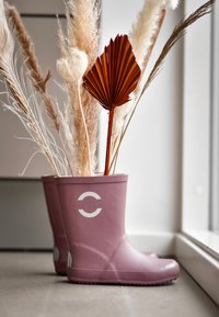 Bottes en caoutchouc violettes remplies de graminées séchées et d'un éventail décoratif orange, présentant un logo blanc sur le côté et une texture lisse et brillante.