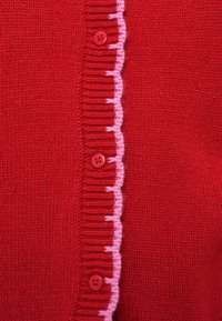 Κόκκινη πλεκτή ζακέτα με κυμματιστή ροζ άκρη κατά μήκος της λωρίδας με κουμπιά, διαθέτει κόκκινα κουμπιά και απαλή υφή.