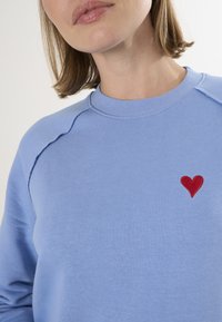 Felpa azzurra con collo rotondo e ricamo a forma di cuore rosso sul lato sinistro. Presenta un tessuto liscio e dettagli di cucitura visibili.
