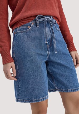 Frau trägt hoch taillierte blaue Denimshorts mit einem gebundenen Gürtel, kombiniert mit einem rostfarbenen Langarmpullover.