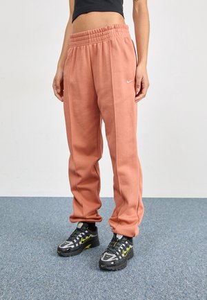 Osoba nosząca łososiowe dresy z białym logo Nike oraz czarne sneakersy Nike z neonowo-żółtymi akcentami, stojąca na niebieskim dywanie.