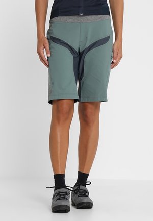 Person wearing green and black knee-length outdoor shorts with black ankle socks and gray hiking shoes against a white background.