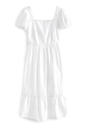 Femme marchant dans une haute herbe près de l'eau au coucher du soleil, portant une robe midi blanche en broderie anglaise et des sandales à lacets.