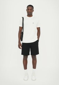 Maglietta di cotone bianca con logo nero, pantaloni corti neri e sneakers bianche con suole testurizzate. Il modello porta una piccola borsa a tracolla nera.