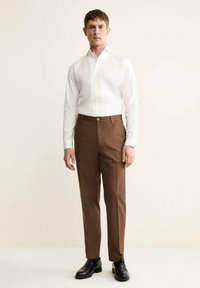 Camicia bianca a maniche lunghe con colletto a punta, abbinata a pantaloni marroni su misura e scarpe in pelle nera. Design semplice e aderente.