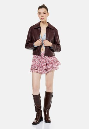 Joven mujer que lleva una chaqueta de cuero marrón oscuro, top azul claro, falda rosa con volantes florales y botas marrón oscuro hasta la rodilla, de pie frente a un fondo blanco.