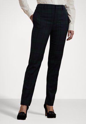 Femme portant un pantalon slim sombre à carreaux avec poches latérales et des chaussures à talons noirs, associé à une chemise blanche à manches longues.