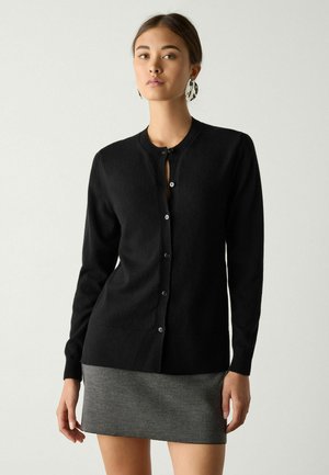 Junge Frau trägt eine schwarze, geknöpfte Strickjacke und einen grauen Minirock, posiert vor einem einfarbigen hellen Hintergrund.