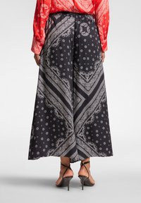 Zwarte wijde broek met een wit paisley- en bloemenpatroon, voorzien van een elastische tailleband en zachte stof, gecombineerd met sandaaltjes met straps.