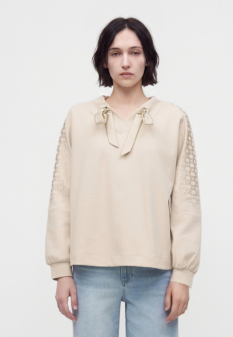Femme aux cheveux courts et foncés portant un haut beige à manches longues avec des découpes décoratives sur les manches et un jean bleu clair, debout devant un fond uni.