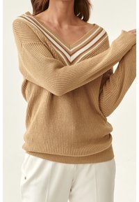 Frau, die einen beigen, gestrickten V-Ausschnitt-Pullover mit weißen und braunen Streifen trägt, kombiniert mit weißen Hosen.