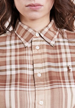 Kobieta w beżowo-brązowej kratkowanej koszuli z kołnierzykiem i zapiętej na guziki, ukazująca usta, podbródek i luźne brązowe włosy.
