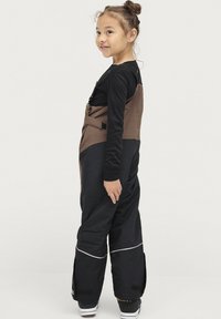 Zwarte en bruine skikleding met verstelbare banden, voorzakken en reflecterende accenten. Comfortabele, losse pasvorm met een gladde textuur.