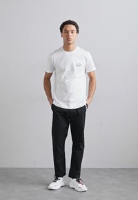 Un tricou alb cu mânecă scurtă, având un mic emblema, asortat cu o pereche de pantaloni negri și adidași albi, pe un fundal neutru.