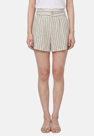 Femme portant un short taille haute beige et blanc à rayures verticales avec deux boutons, un haut blanc et des sandales beiges, debout devant un fond uni.