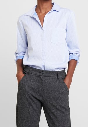 Person wearing a light blue button-up shirt with rolled sleeves tucked into dark gray speckled trousers, hands in pockets.