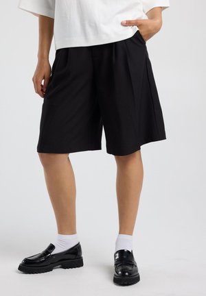 Persoon draagt zwarte knielange shorts, witte sokken, zwarte loafers en een wit shirt, met één hand in de zak, staand binnenshuis.