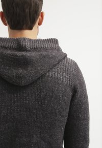 Mörkgrå stickad tröja med huva, med ett texturerat mönster på axlarna och mjukt, grovt tyg.