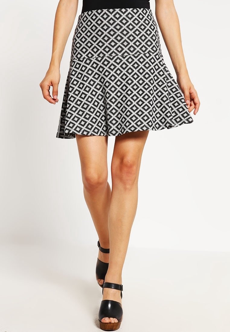 Jupe évasée noire et blanche à motif losanges portée avec des talons noirs ouverts à la cheville, jambes visibles de mi-cuisse jusqu'en bas.
