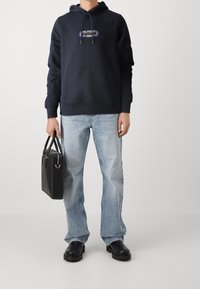 Uomo con felpa blu scuro, jeans azzurri, stivali neri che tiene una valigetta in pelle nera contro uno sfondo semplice.