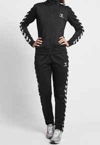 Woman wearing a black Hummel tracksuit with white chevron stripes on sleeves and pants, standing with hands on hips.