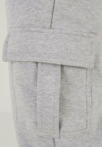 Grijze sweatshirtstof met een detail van een opgestikte zak met klep. De textuur lijkt zacht en heeft een subtiel gemêleerd patroon.