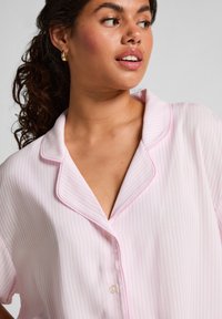 Vrouw met krullend donker haar in een roze en witte gestreepte button-up shirt die naar de zijkant kijkt tegen een effen achtergrond.