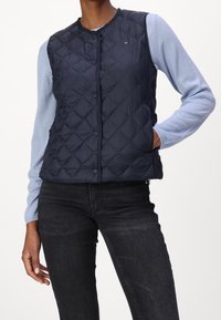 Marineblau gesteppte Pufferweste mit Druckknöpfen, Rundhalsausschnitt und leichtem Material, über einem hellblauen Langarmshirt getragen.