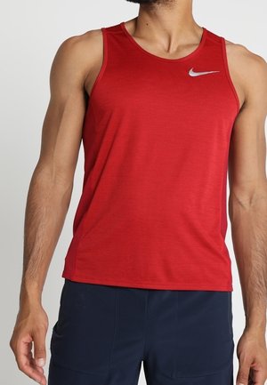 Homme portant un haut de sport rouge sans manches avec un petit logo blanc Nike sur le côté gauche de la poitrine et un short bleu foncé sur un fond uni.