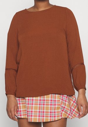 Osoba ubrana w top z długim rękawem koloru rdzawego z koronkowymi detalami na rękawach oraz beżową, czerwoną i białą plisowaną spódnicę.