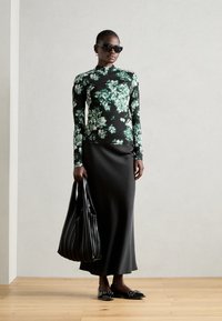 Langarm bloemtop in zwart en groen, gecombineerd met een zwarte, aansluitende maxirok. Accessoires omvatten zwarte zonnebril en een geplooide tas.
