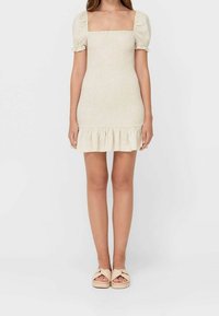 Robe courte beige avec un décolleté carré, des manches bouffantes et un ourlet à volants. Fabriquée en tissu texturé. Associée à des sandales beige nouées.