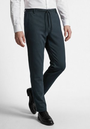 Man draagt donkere slim-fit broek met trekkoord in de taille, wit overhemd en zwarte leren nette schoenen, staand met één been licht gebogen.