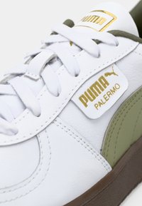 Zapatilla de cuero blanca con acentos en verde oliva, con el logo de Puma en dorado y blanco, cordones blancos planos y suela de goma marrón.