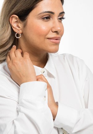 Orecchini a cerchio in oro rosa con piccoli strass trasparenti, che presentano un design circolare con superficie texturizzata, indossati con una camicia bianca a bottoni.