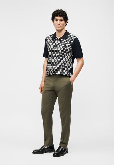 Νεαρός άνδρας στέκεται με το ένα χέρι στην τσέπη, φοράει κοντομάνικο πόλο με ασπρόμαυρο γεωμετρικό σχέδιο, ελαιδί παντελόνι και μαύρα παπούτσια.
